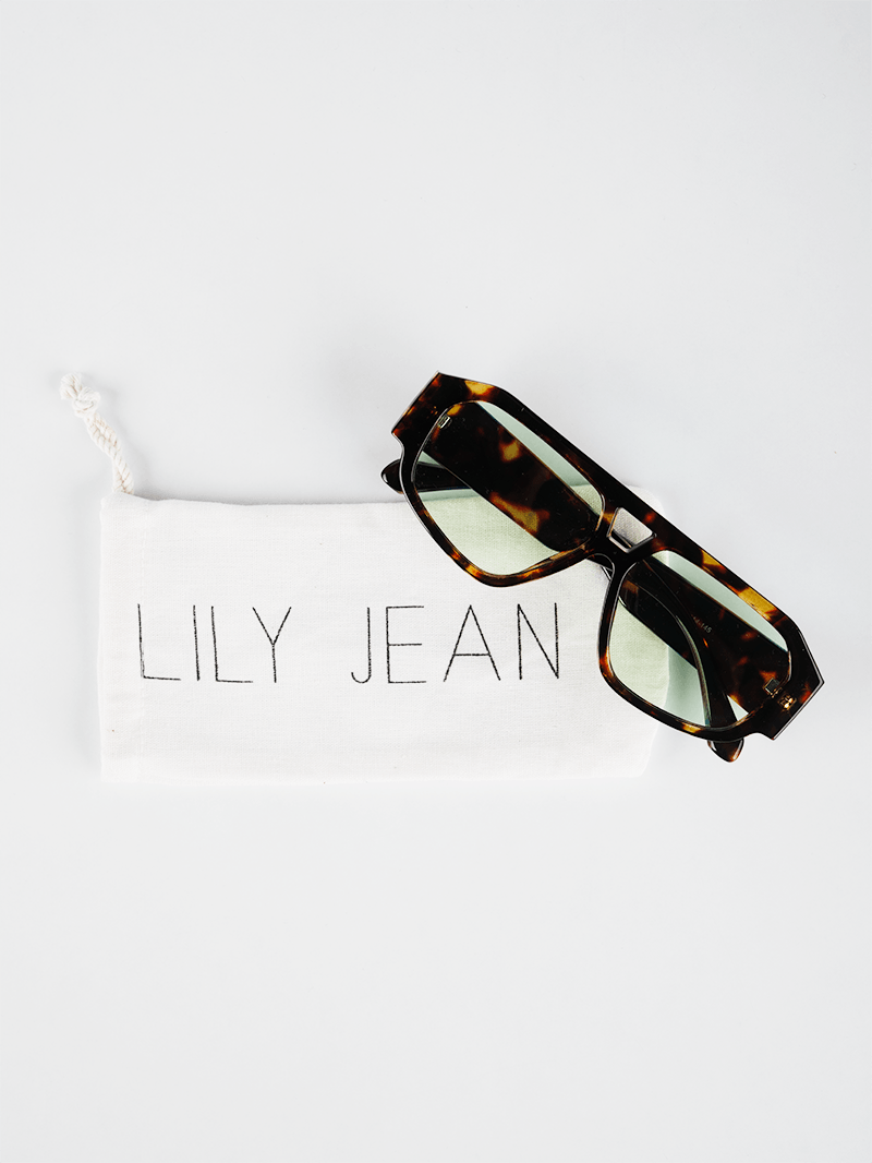 Oversized rectangle sunglasses with a dark brown and amber tortoiseshell frame and tinted light green lenses on a white drawstring packaging pouch by Lily Jean