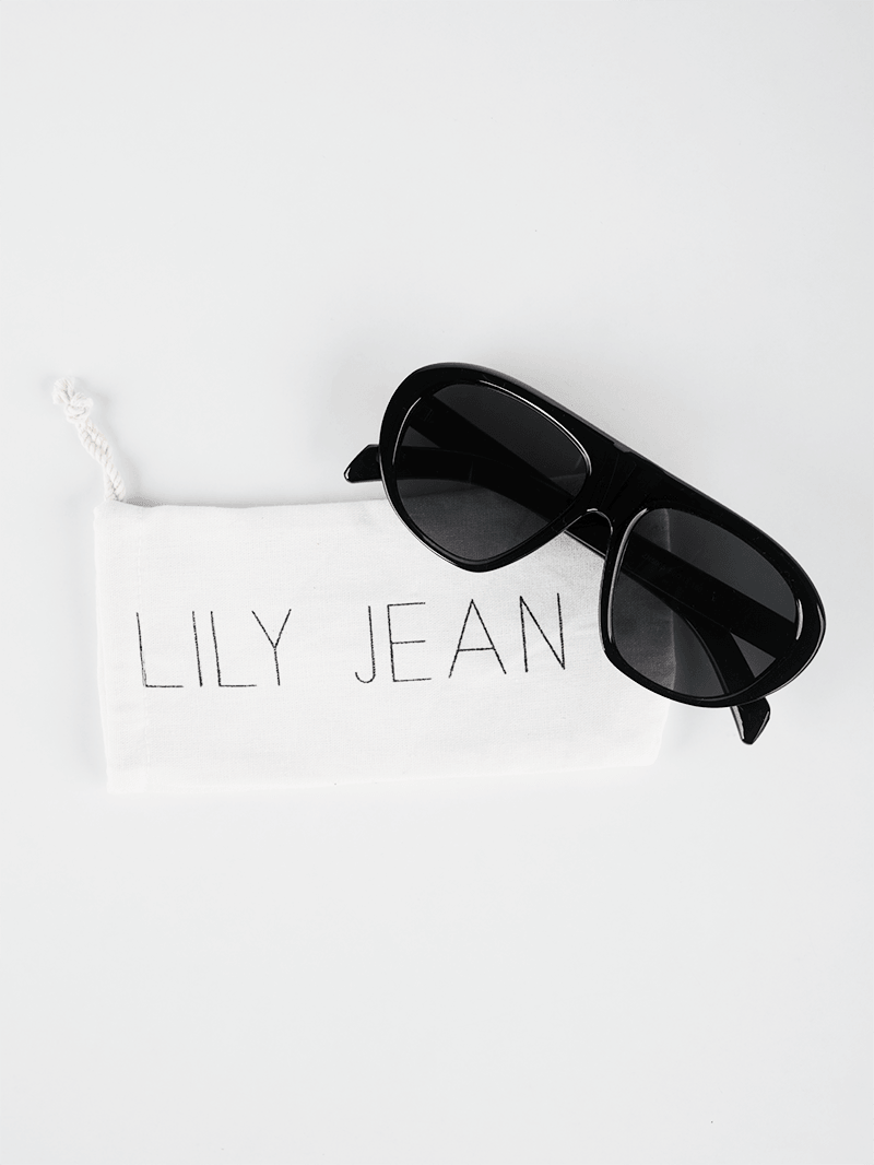 Square-aviator black sunglasses with dark gray lenses on a white drawstring packaging pouch by Lily Jean