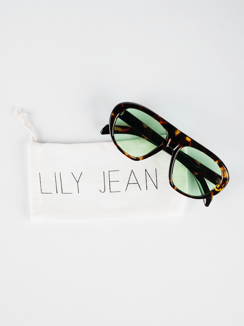 Square-aviator tortoise shell sunglasses with tinted green lenses on a white drawstring packaging pouch by Lily Jean